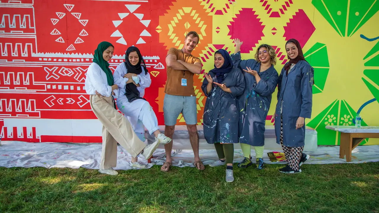 Group of people smiling and posing with their hands and feet in the air, in front of a colorful, hand-painted mural